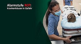 Das Bild zeigt eine medizinische Fachkraft, die eine Patientin auf einer Trage in ein Krankenhaus schiebt. Im Hintergrund wird die Alarmstufe Rot angezeigt, was auf eine gefährliche Situation für die Krankenhäuser hinweist.