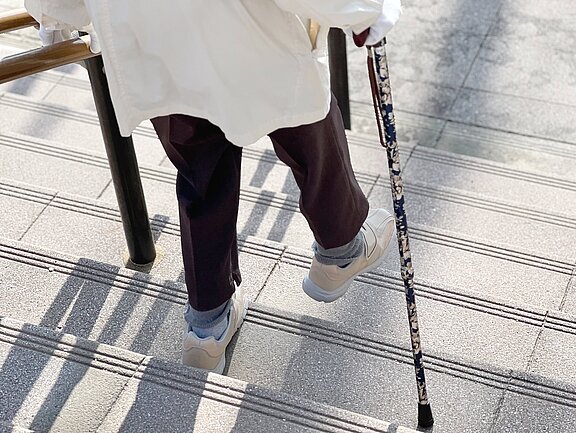 Eine Person mit einer weißen Jacke und dunklen Hosen geht eine Treppe hinunter, während sie sich mit einem blumigen Gehstock abstützt. Ihre Schuhe sind hell und sie hat graue Socken an. Die Stufen sind mit Rillen versehen.