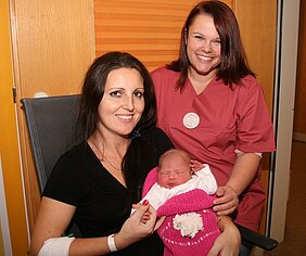 Eine lächelnde Frau hält ein Neugeborenes, das in eine rosa Decke gewickelt ist. Neben ihr steht eine Krankenschwester ebenfalls lächelnd. Der Hintergrund zeigt eine Krankenhausumgebung.