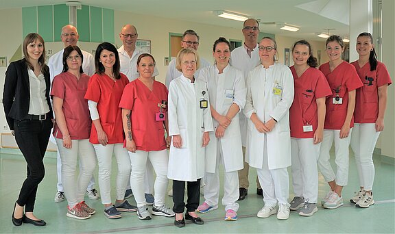 Eine Gruppe von 15 Personen steht in einem Krankenhausflur. Die Mehrheit trägt rote oder weiße Arbeitskleidung, einige in Laborkitteln. Alle lächeln freundlich in die Kamera, während sie eng zusammenstehen. Im Hintergrund sind die Wände des Flurs sichtbar.