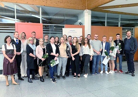 Eine Gruppe von 20 Personen steht in einem Raum, einige halten Blumen und Urkunden in der Hand. Im Hintergrund sind Wandpanels sichtbar. Die Atmosphäre wirkt festlich und feierlich. Eine Person ist in traditioneller Tracht gekleidet.