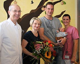 Eine Familie steht zusammen in einem Krankenhaus. Der Vater hält ein neugeborenes Baby, während die Mutter einen Blumenstrauß hält. Zwei Pflegekräfte lächeln freundlich neben ihnen. Im Hintergrund ist eine Wand mit einem Baum und Äpfeln zu sehen.