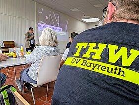 Eine Person steht vor einer Gruppe, die in einem Raum sitzt. Auf einem Bildschirm im Hintergrund steht „Herzlich Willkommen“ und ein Thema zum Katastrophenschutz. Im Vordergrund ist das Rückenprofil eines Teilnehmers zu sehen, der ein T-Shirt mit der Aufschrift „THW OV Bayreuth“ trägt.