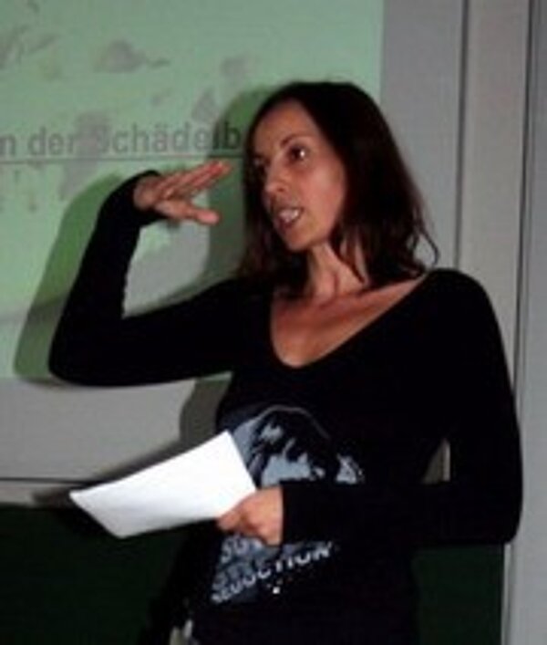 Eine Frau mit langen, braunen Haaren steht an einem Rednerpult. Sie hält ein Blatt Papier in der Hand und gestikuliert mit der anderen Hand. Im Hintergrund sind schemenhaft Präsentationsfolien sichtbar.