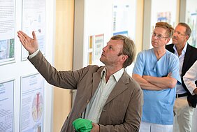 Ein Mann in einem hellen Sakko erklärt eine Präsentation an einer Wand. Er hält ein grünes Tuch in der Hand und zeigt mit der anderen Hand auf die Informationen. Im Hintergrund stehen mehrere Personen, die aufmerksam zuhören.