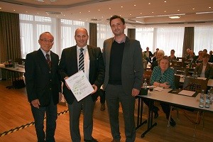 Drei Männer stehen in einem Konferenzraum zusammen. Der Mann in der Mitte hält ein Dokument in den Händen. Im Hintergrund sind Tische mit Teilnehmern, die an einer Besprechung teilnehmen. Die Atmosphäre wirkt formell und professionell.