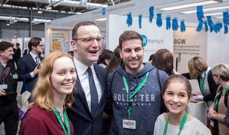 Vier Personen lächeln in einer Messe-Umgebung. Ein Mann in Anzug steht in der Mitte und posiert mit zwei jungen Frauen und einem jungen Mann. Alle tragen Namensschilder und stehen vor Ständen, die teilweise nicht sichtbar sind. Die Atmosphäre wirkt freundlich und entspannt.