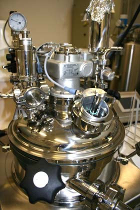 Ein hochglänzender, runder Druckbehälter aus Edelstahl mit verschiedenen Ventilen, Druckmanometern und Rohren. Die komplexe Anordnung von mechanischen Teilen deutet auf eine technische Anwendung hin, möglicherweise in einem Labor oder industriellen Umfeld.