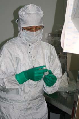 Eine Person in einem weißen Schutzanzug und grünen Handschuhen hält ein kleines Behältnis in der Hand. Die Person trägt eine Schutzmaske und eine Schutzbrille und arbeitet in einem Labor oder einer ähnlichen Umgebung.