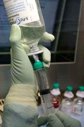 Eine Hand mit grünen Handschuhen hält eine Spritze, die mit Flüssigkeit gefüllt wird. Die Spritze hat eine grüne Kappe und eine Skala, die den Füllstand anzeigt. Im Hintergrund sind mehrere Fläschchen zu sehen, die medizinische Substanzen enthalten.