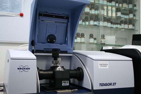 Das Bild zeigt ein wissenschaftliches Gerät, das als Fourier-Transformations-Infrarotspektrometer dient. Es hat eine kastenförmige Bauweise mit einem offenen Deckel und einem zentralen Halter für Proben. Im Hintergrund sind Regale mit Fläschchen sichtbar, die verschiedene chemische Substanzen enthalten.