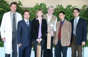 Sechs Personen stehen in einer Gruppe, umgeben von Pflanzen. Sie tragen formelle Kleidung und zeigen verschiedene Ausdrücke. In der Mitte hält eine Frau eine Tasche. Die Szene vermittelt eine Atmosphäre von Zusammenarbeit oder Feierlichkeit.