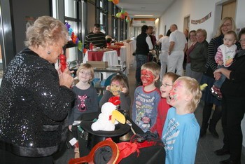 Eine ältere Frau mit glitzerndem Oberteil unterhält eine Gruppe von Kindern, die mit Gesichtern bemalt sind. Die Kinder schauen gespannt zu, während die Frau einen Zauberhut und Requisiten präsentiert. Im Hintergrund sind weitere Personen und festliche Dekorationen zu sehen.