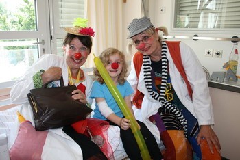 Zwei Clowns in weißen Kostümen und mit roten Nasen posieren mit einem Kind, das einen grünen Ballon in der Hand hält. Alle drei lächeln und scheinen eine fröhliche Stimmung zu haben. Im Hintergrund sind helle Wände und Fenster sichtbar.