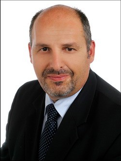 Ein Mann mit kurzen, grauen Haaren und einem gepflegten Bart lächelt in die Kamera. Er trägt einen schwarzen Anzug und ein hellblaues Hemd mit dunkler Krawatte. Der Hintergrund ist neutral und hell.