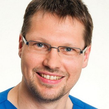 Ein lächelnder Mann mit Brille trägt ein blaues Oberteil. Sein Gesicht zeigt ein freundliches und offenes Ausdruck. Der Hintergrund ist neutral und hell gestaltet.