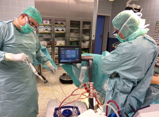 Zwei medizinische Fachkräfte in grünen OP-Kitteln und Schutzmasken arbeiten miteinander in einem Behandlungsraum. Ein Monitor zeigt medizinische Daten an, während einer der Fachkräfte eine Spritze in ein Gerät einführt. Der Raum enthält weitere medizinische Geräte und ist ordentlich organisiert.