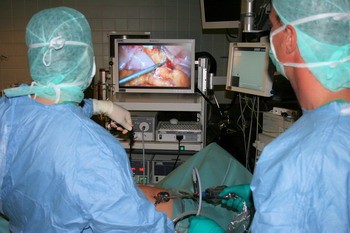 Zwei Chirurgen in OP-Kleidung führen eine minimalinvasive Operation durch. Sie schauen auf einen Bildschirm, der das Operationsfeld zeigt. In ihren Händen halten sie chirurgische Instrumente, während sie mit einem Endoskop arbeiten. Der Fokus liegt auf der präzisen Durchführung des Eingriffs.