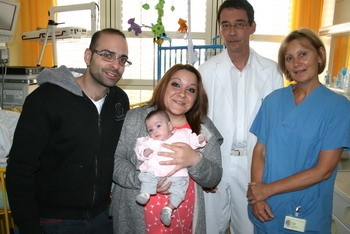 Eine Familie mit einem Neugeborenen steht lächelnd in einem Krankenhauszimmer. Die Mutter hält das Baby, während der Vater neben ihr steht. Hinter ihnen stehen ein Arzt in einem weißen Kittel und eine Krankenschwester in blauer Kleidung. Die Atmosphäre ist freundlich und unterstützend.