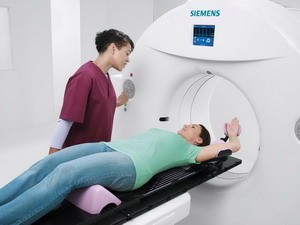 Eine medizinische Fachkraft steht neben einem Patienten, der auf einer Liege liegt und sich in einer MRT-Maschine befindet. Der Patient trägt ein grünes T-Shirt und hat die Arme seitlich angelegt. Die Fachkraft spricht mit dem Patienten, während das Gerät bereit ist, zu arbeiten.