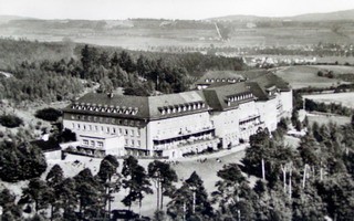 Ein historisches Foto eines großen Gebäudes, möglicherweise eines Hotels oder einer Kurklinik, umgeben von Bäumen und einer hügeligen Landschaft. Das Bild vermittelt eine ruhige, ländliche Atmosphäre.