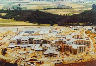 Luftaufnahme einer Baustelle mit mehreren Gebäuden in verschiedenen Bauphasen. Umgeben von Feldern und Wäldern, sind Kräne und Baustellenfahrzeuge sichtbar. Die Baustelle zeigt die Entwicklung eines größeren Gebäudekomplexes.