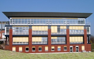 Ein modernes, dreigeschossiges Gebäude mit großen Fenstern und einem flachen Dach. Die Fassade ist rot mit gelben Jalousien und klaren Gebäudestrukturen. Der Himmel ist blau und sonnig, was dem Gebäude ein einladendes Aussehen verleiht.