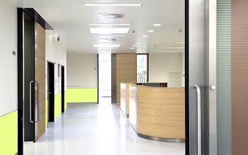 Ein moderner Empfangsbereich mit Holz- und Glaselementen. Links sind zwei Türen sichtbar, eine geschlossene und eine mit einem transparenten Fenster. Der Empfangstresen aus Holz befindet sich im Hintergrund, und die Wände sind unten in einem hellen Gelb gestrichen.